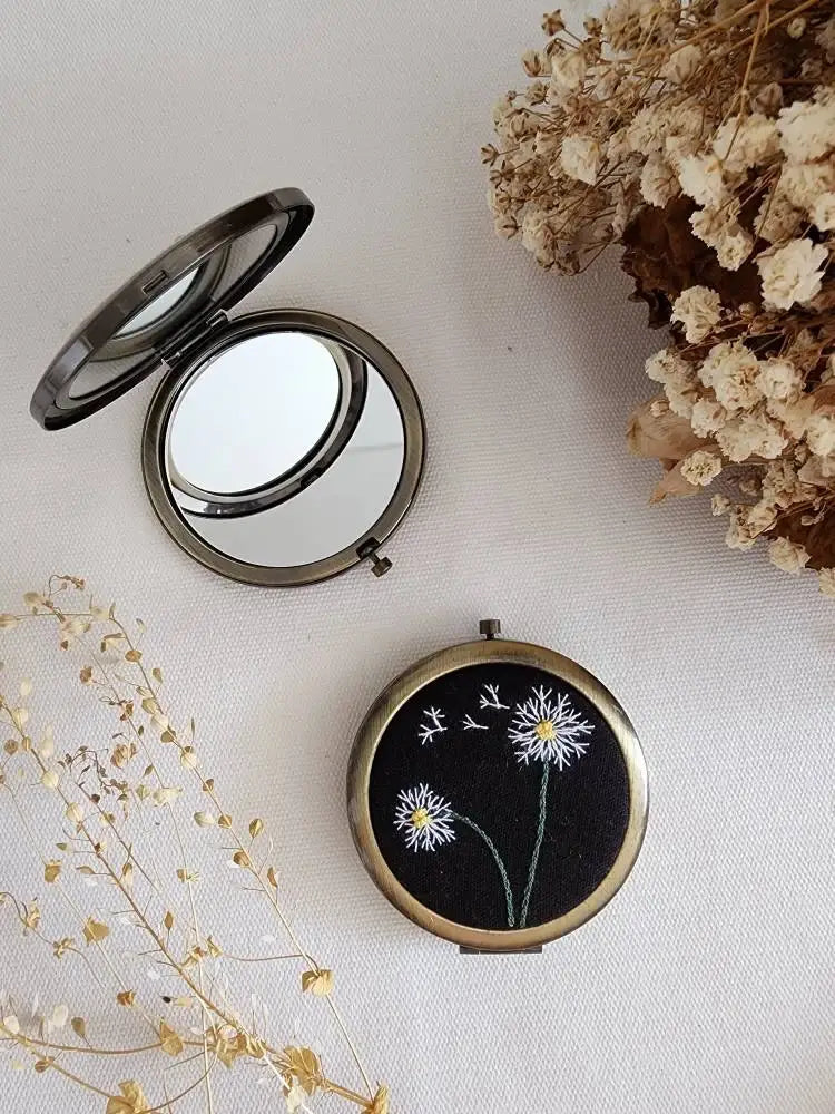 Compact mirror with floral design on a light surface with dried flowers.