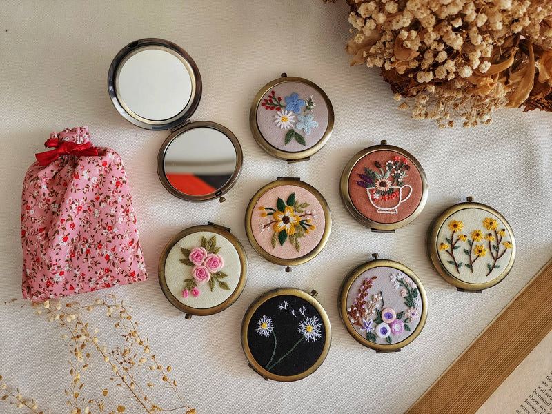 Set of compact mirrors with floral designs on a light surface with a pink floral bag and dried flowers.