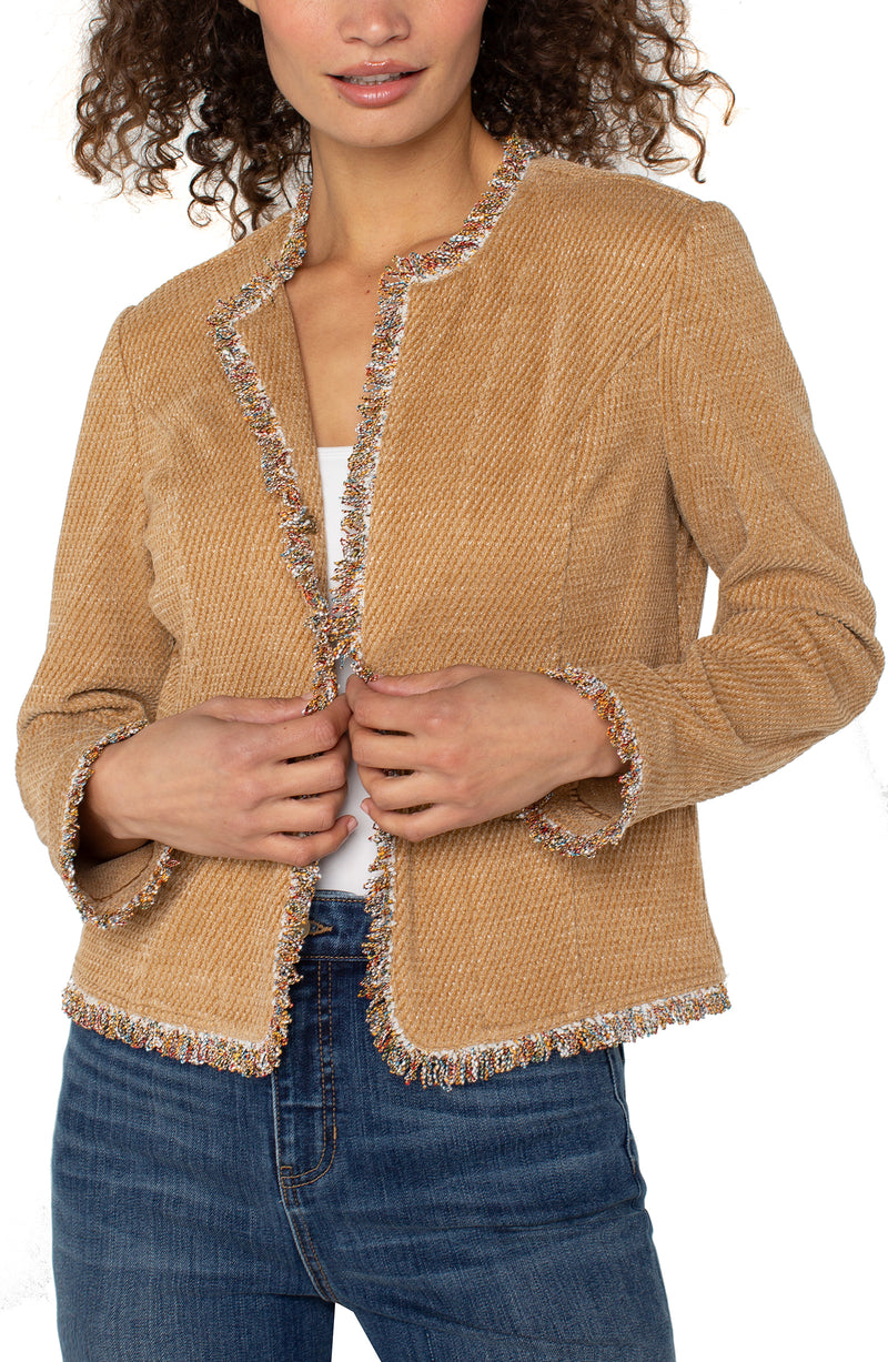 A woman wearing a honey-colored collarless jacket with frayed edges over a white top, paired with blue jeans.